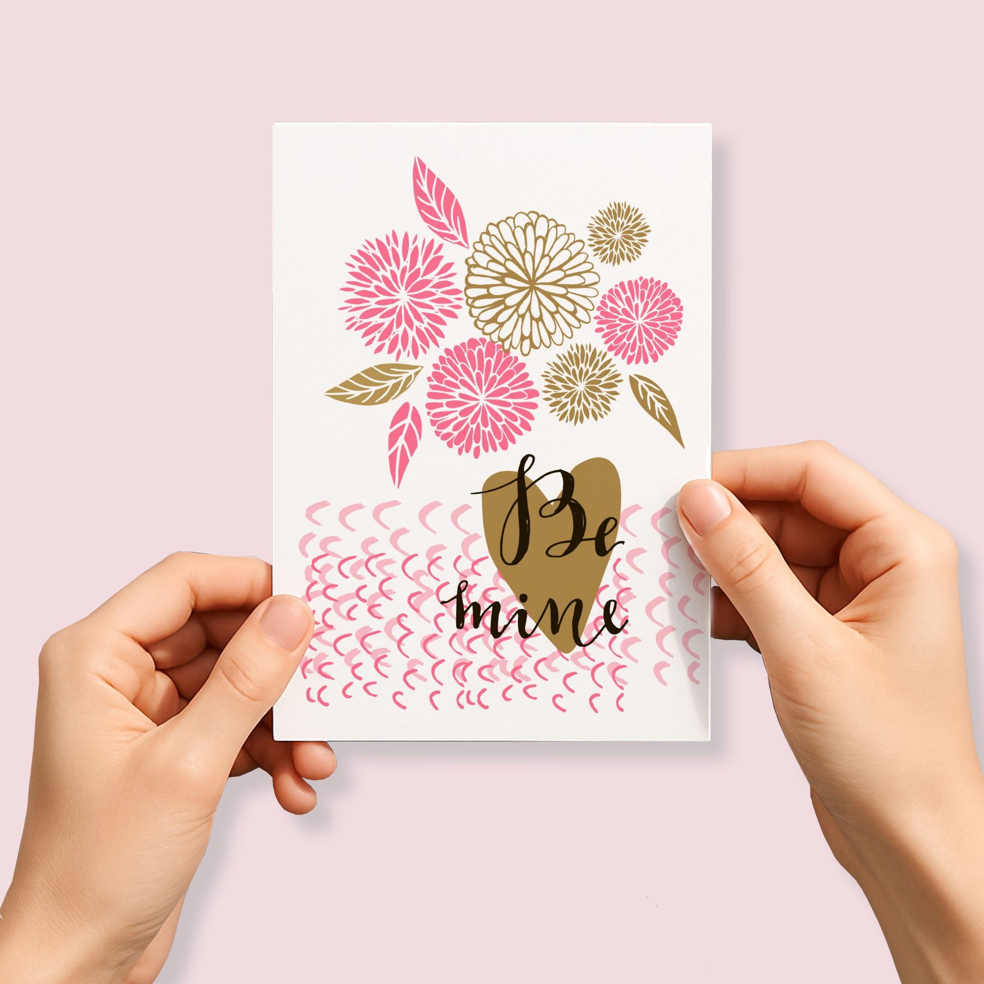 Kjærlighetskort med teksten Be mine, dekorative blomster, blader og håndtegnet hjerte i rosa og gylne toner. Person som holder kortet.