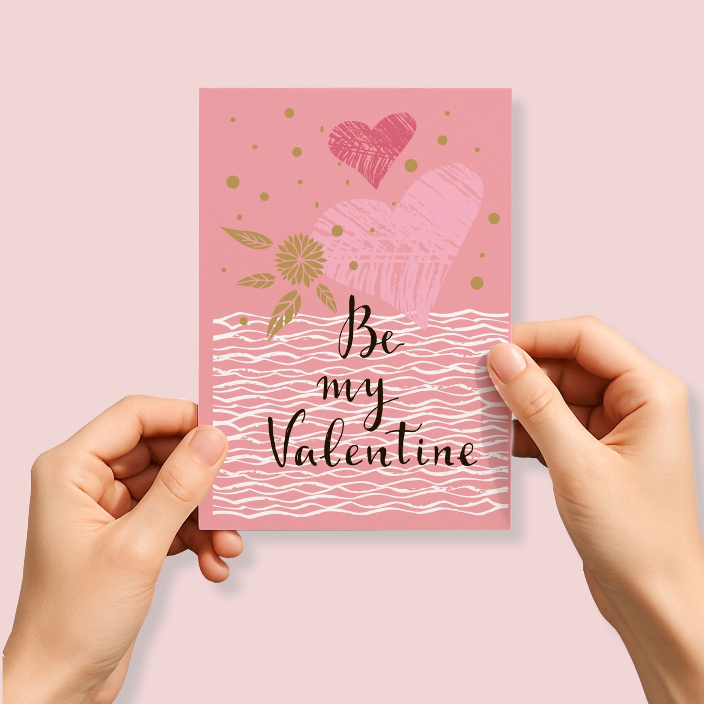 Kjærlighetskort med teksten Be my Valentine, håndtegnede hjerter, bølgende linjer og dekorative detaljer i rosa og gylne toner. Person som holder kortet.