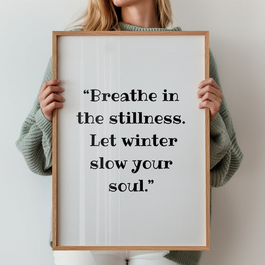 Kvinne holder innrammet plakat med teksten “Breathe in the stillness. Let winter slow your soul.” i et lyst, nordisk interiør.