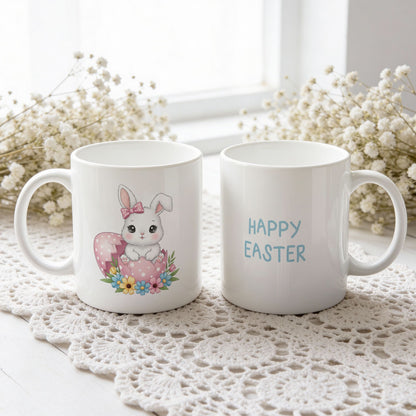 Påskekrus med hvit kanin med rosa sløyfe i rosa egg og teksten Happy Easter, vist fra begge sider på hvit blondeduk.