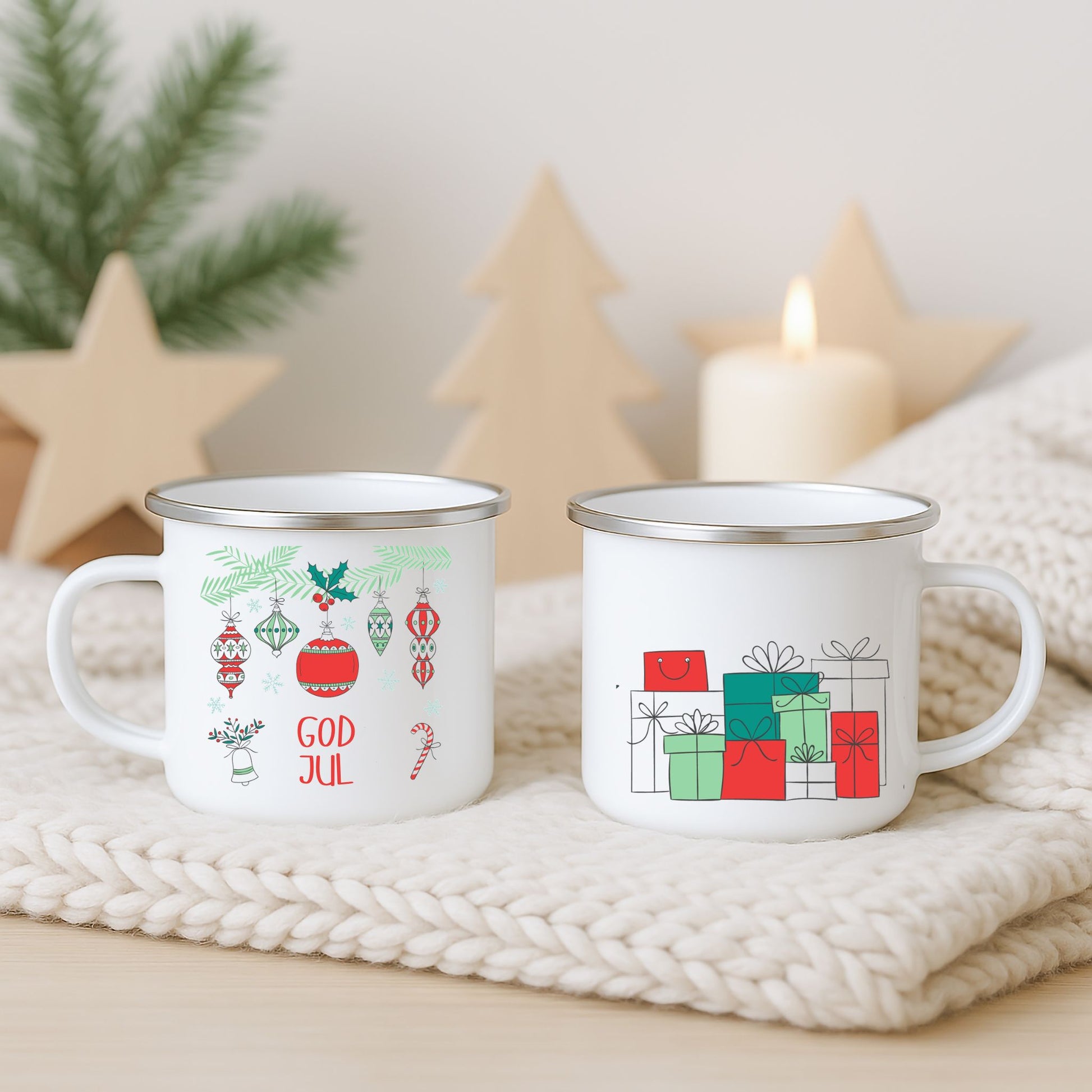 Hvitt emaljekrus med to grafiske julemotiver: hengende ornamenter med teksten «God Jul» og en minimalistiske julegaver i rødt og grønt, fotografert på lys strikket duk med juledekor i bakgrunnen.