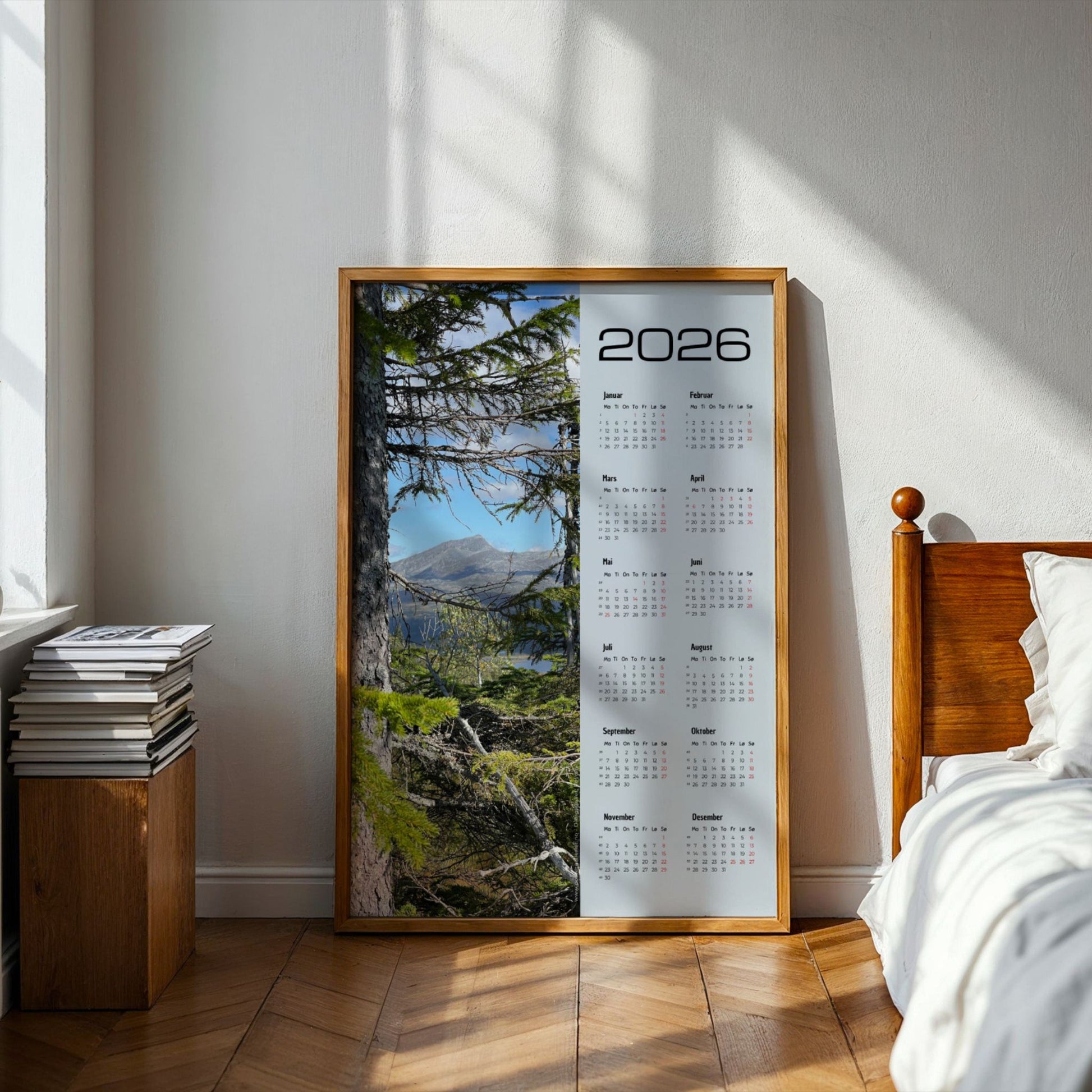 Kalender 2026 med fjellmotiv sett mellom kvister fra et stort grantre i Røyrvik. Illustrasjon viser kalender i eik ramme på et soverom.