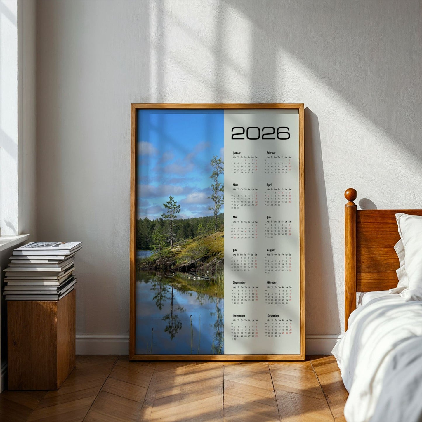Fotokalender 2026 med landskapsmotiv fra Midterfjellet i Steinkjer, stille vann som speiler skog og himmel, med norsk kalender og røde dager. Illustrasjon viser kalender i eik ramme på et soverom.