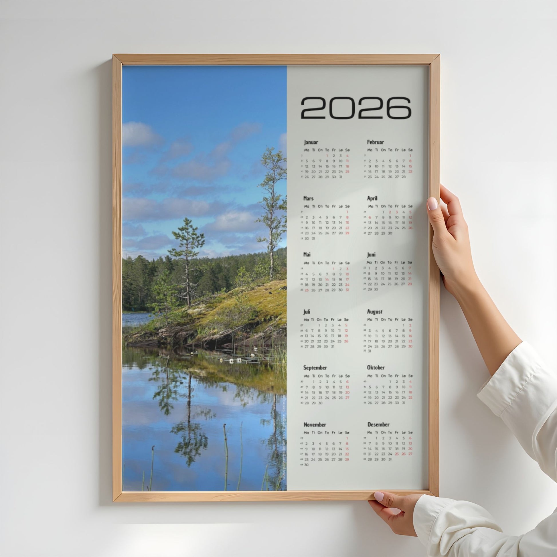 Fotokalender 2026 med landskapsmotiv fra Midterfjellet i Steinkjer, stille vann som speiler skog og himmel, med norsk kalender og røde dager. Illustrasjon viser kalender i eik ramme.
