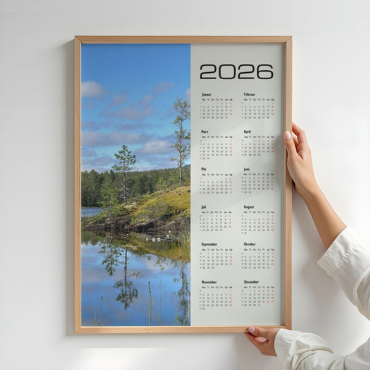 Fotokalender 2026 med landskapsmotiv fra Midterfjellet i Steinkjer, stille vann som speiler skog og himmel, med norsk kalender og røde dager. Illustrasjon viser kalender i eik ramme.