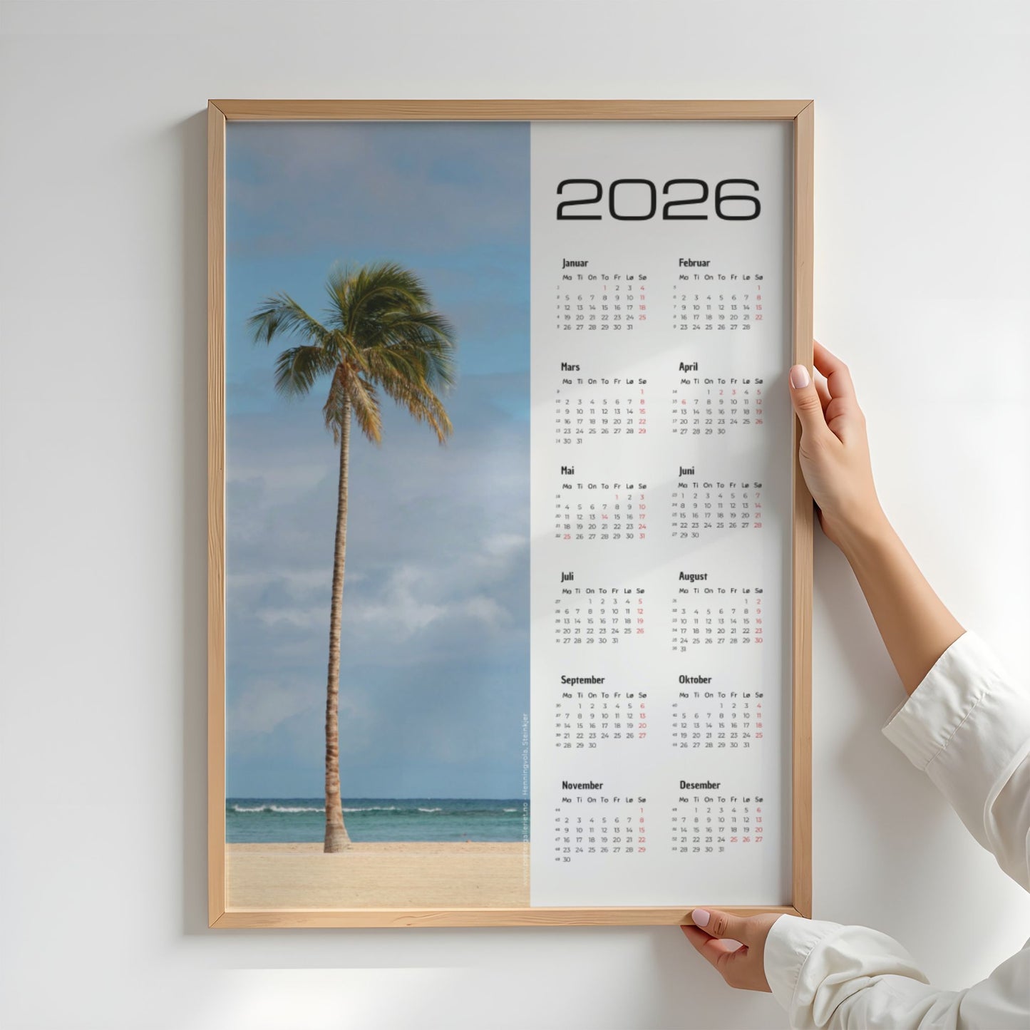 Kalender 2026 med motiv av en enslig palme på en rolig tropisk strand, med oversiktlig kalenderdel og norske merkedager. Illustrasjon viser kalender i eik ramme.