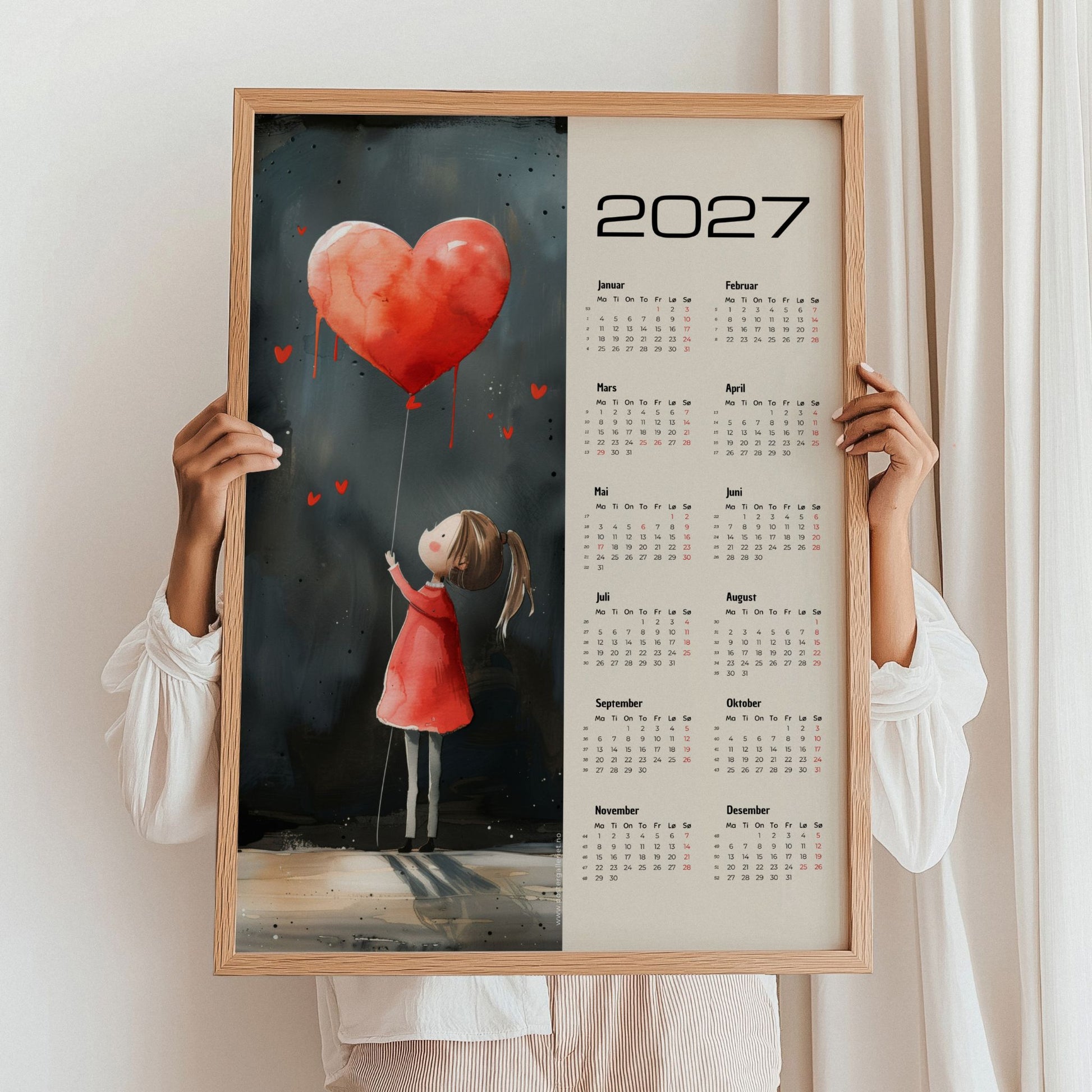 Kalender 2027 med pike, rød hjerteballong og beige bakgrunn, holdt i eikeramme.