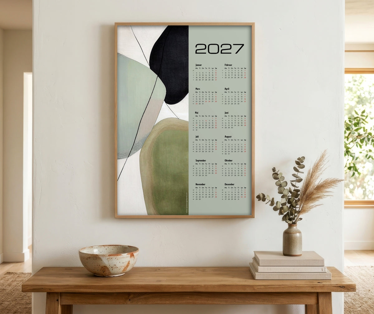 Grafisk veggkalender 2027 med abstrakte former i grønne, beige og sorte toner, innrammet over et trebord i en lys, nordisk entré.