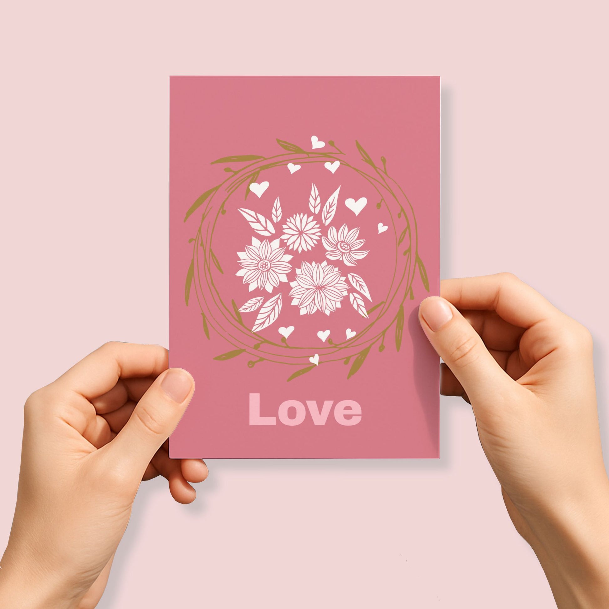 Kjærlighetskort med teksten Love, dekorativ blomsterkrans, blomster og små hjerter i hvitt og gylne toner på rosa bakgrunn. Person som holder kortet.