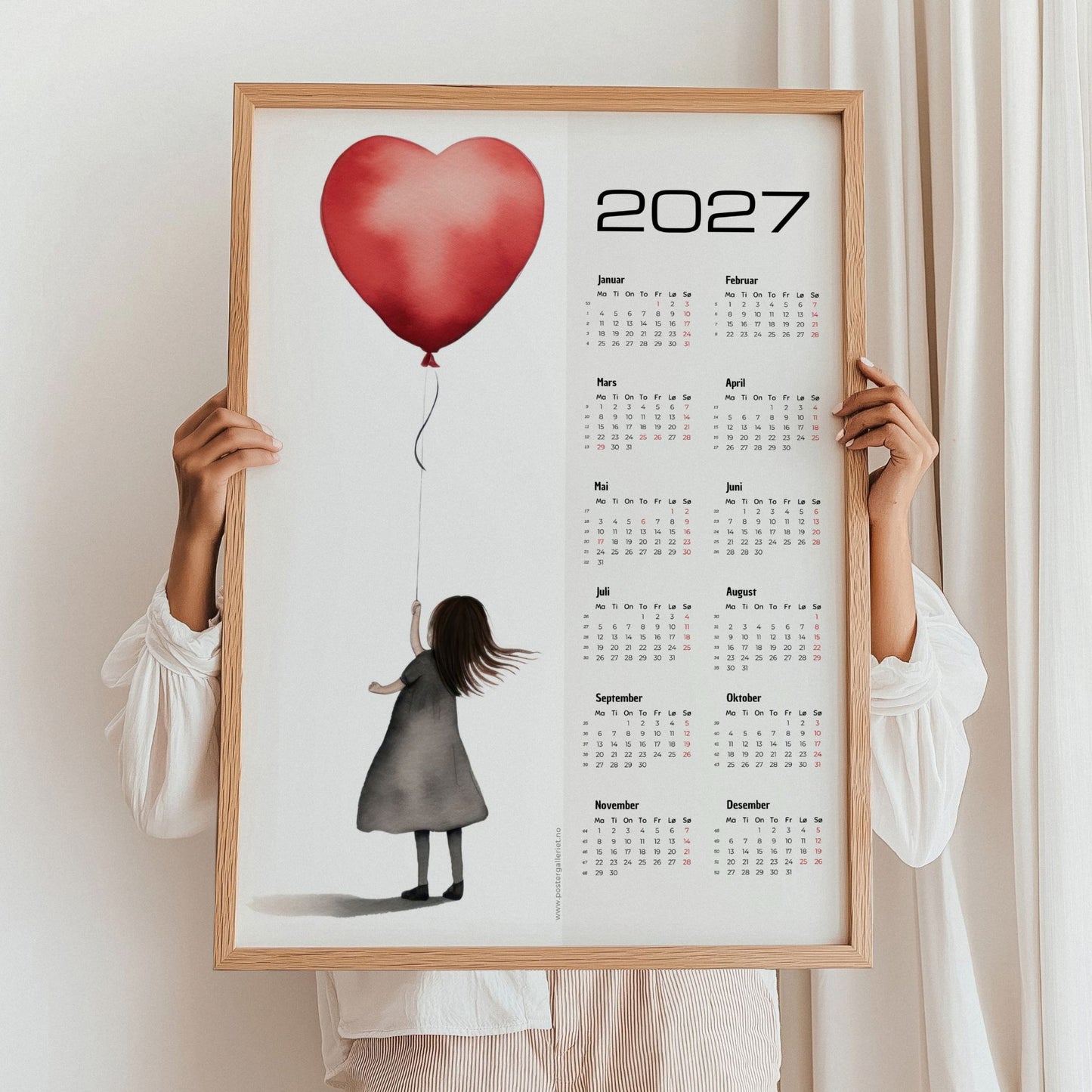 Minimalistisk kalender 2027 med jente og rød hjerteballong, holdt opp i eikeramme.