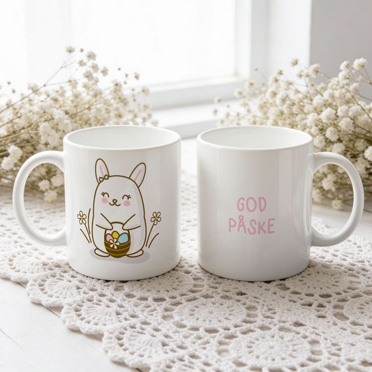 Hvitt keramikkrus med smilende påskehare omgitt av blomster og kurv med påskeegg, samt teksten God påske i rosa tone.
