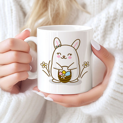 Hvitt keramikkrus med smilende påskehare omgitt av blomster og kurv med påskeegg, samt teksten God påske i rosa tone. Person holder kruset.