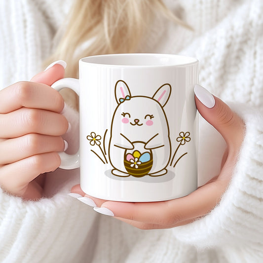 Hvitt keramikkrus med smilende påskehare omgitt av blomster og kurv med påskeegg, samt teksten God påske i rosa tone. Person holder kruset.