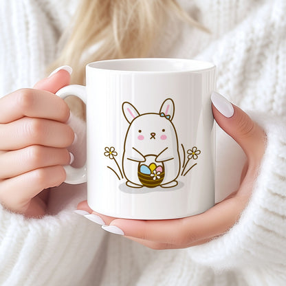 Hvitt keramikkrus med søt påskehare med sløyfe og kurv med påskeegg, samt teksten God påske i blå tone på motsatt side. Person holder kruset.