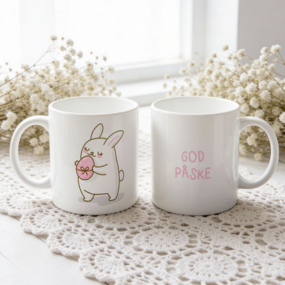 Hvitt keramikkrus med søt påskehare som holder rosa påskeegg og teksten God påske i rosatone på motsatt side.