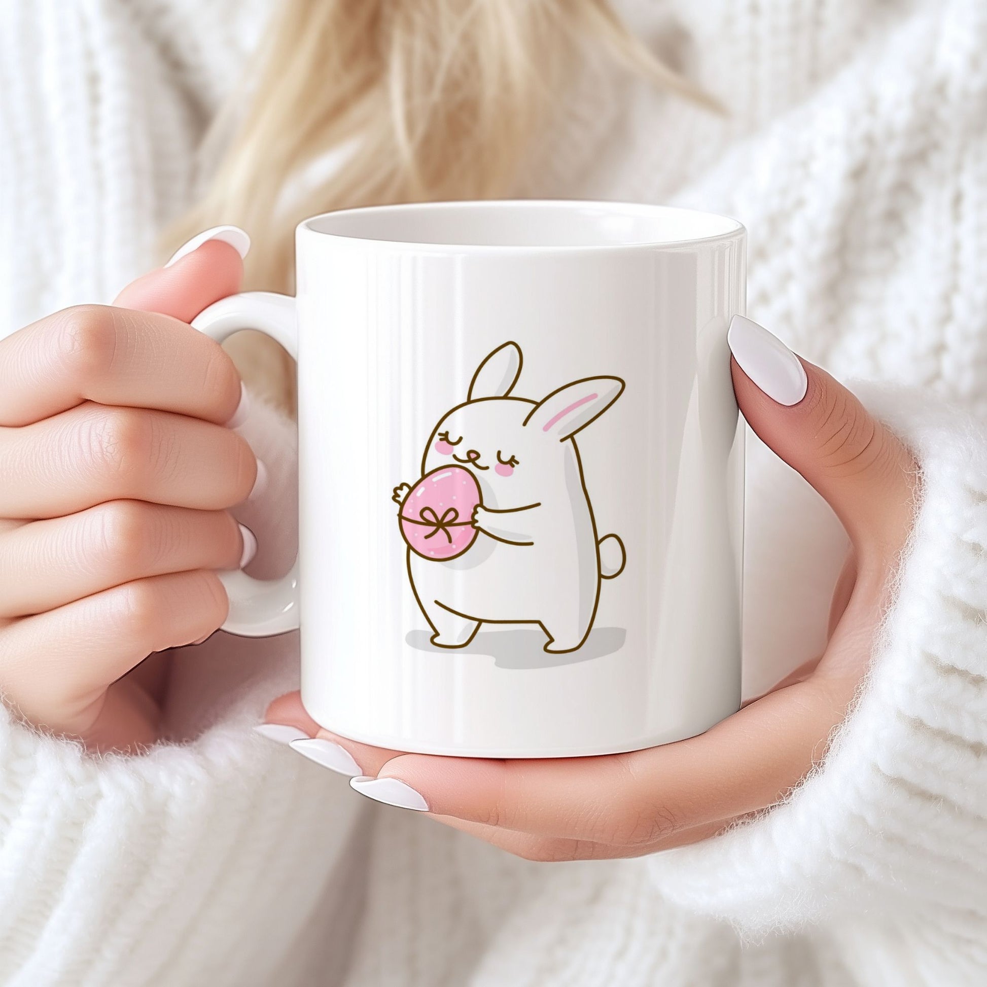 Hvitt keramikkrus med søt påskehare som holder rosa påskeegg og teksten God påske i rosatone på motsatt side. Person holder krus.