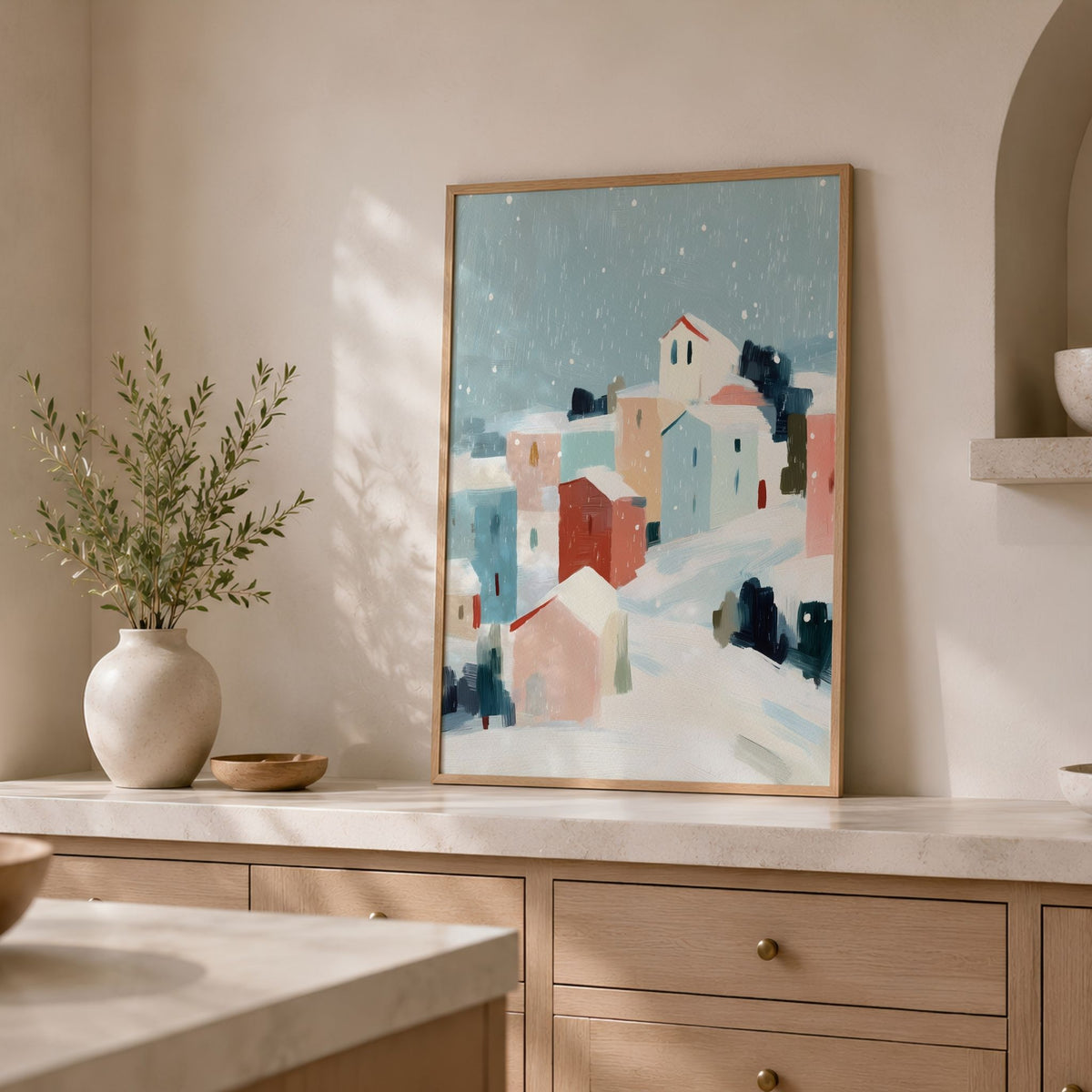 Snowfall Village plakat i eikeramme på lys kjøkkenbenk med grønne grener i vase.