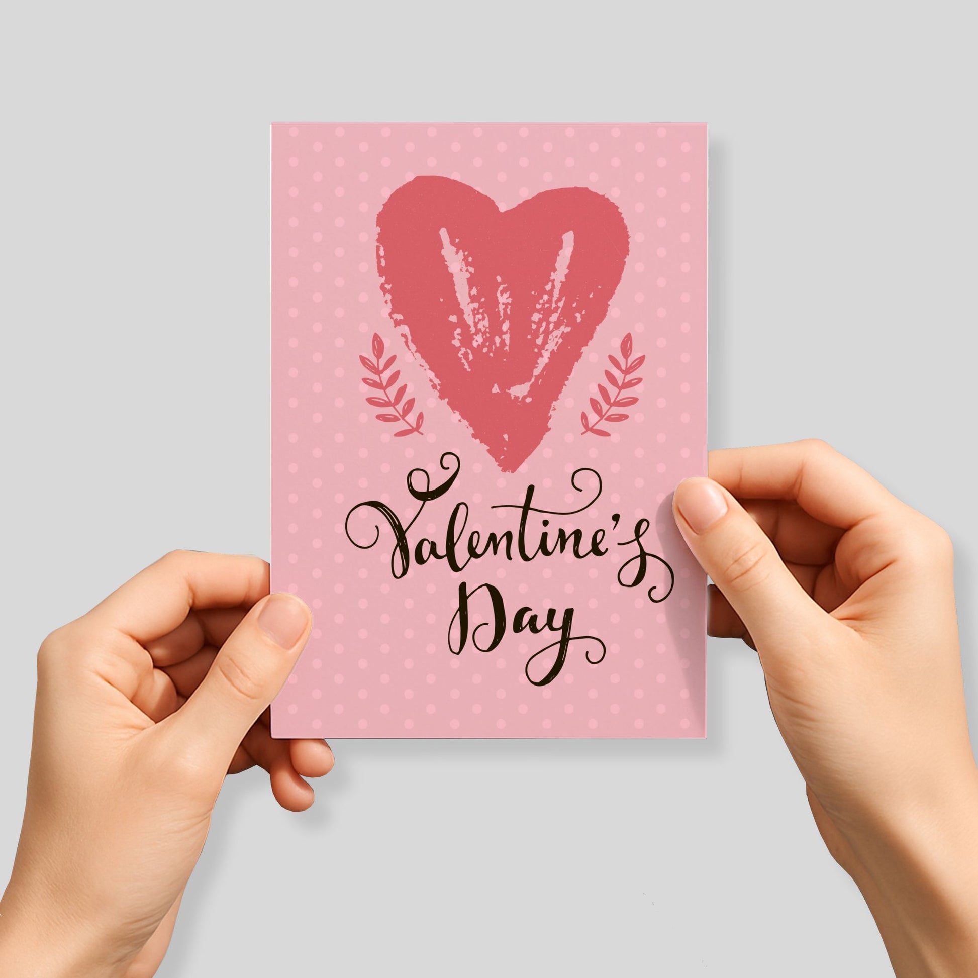 Kjærlighetskort med teksten Valentine’s Day, stort håndmalt hjerte og rosa prikket bakgrunn med botaniske detaljer. Person som holder kortet.