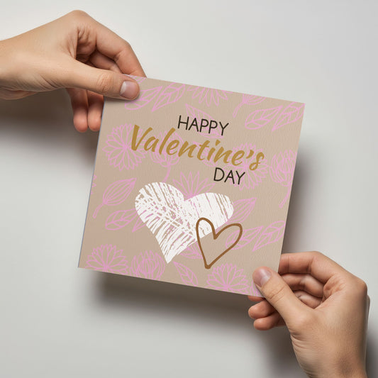 Valentineskort med teksten Happy Valentine’s Day, håndtegnede hjerter og floralt mønster i beige og rosa toner. Kortet blir holdt av en person.